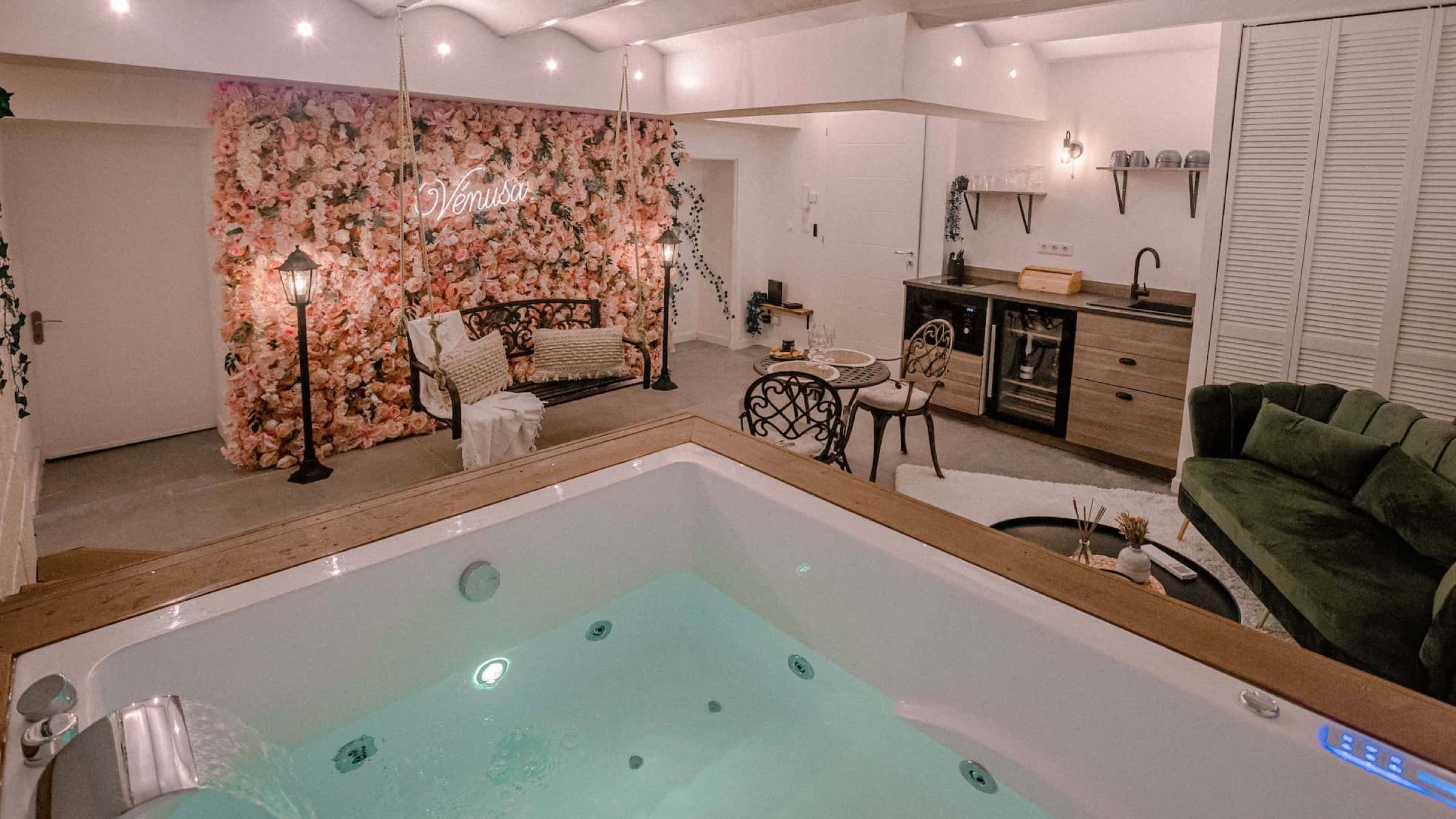 Jacuzzi en eau au premier plan, mur fleuri, balançoire et kitchenette en arrière-plan.